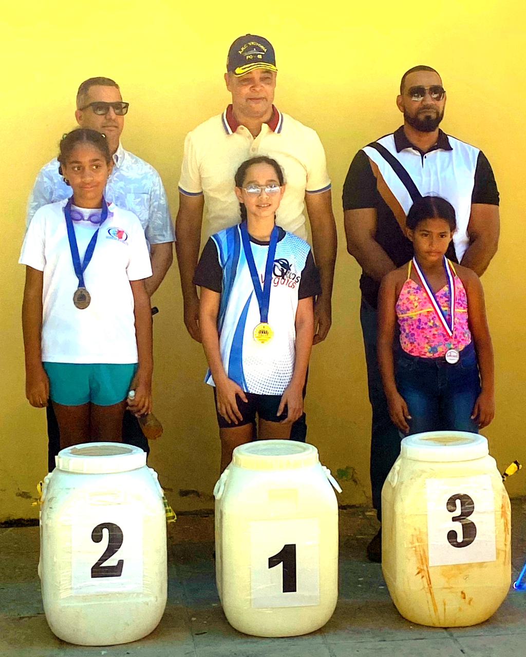 Camille Estévez (oro), Nicole Quezada (plata) y Kiara González (bronce), son premiadas por José Luis Bautista, José R. Muñoz Monción y Octavio Martínez.