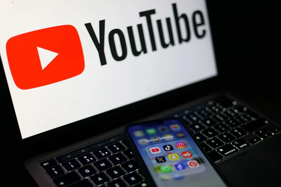 Pantalla con el logo de YouTube, propiedad de Alphabet, y un móvil con aplicaciones de redes sociales como Instagram y Facebook, de Meta.