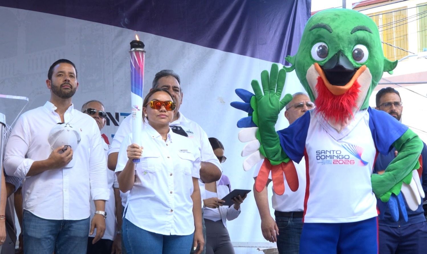 La gobernadora de Moca, Patricia Rivera, sostiene la antorcha de los Juegos Santo Domingo 2026, mientras Colí, la mascota oficial, saluda al público.